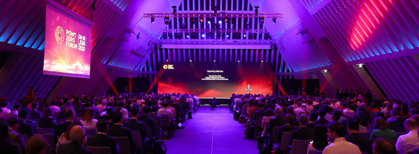 Audience at Point Zero Forum. Speaker on stage with large screen. Purple and red stage lighting.